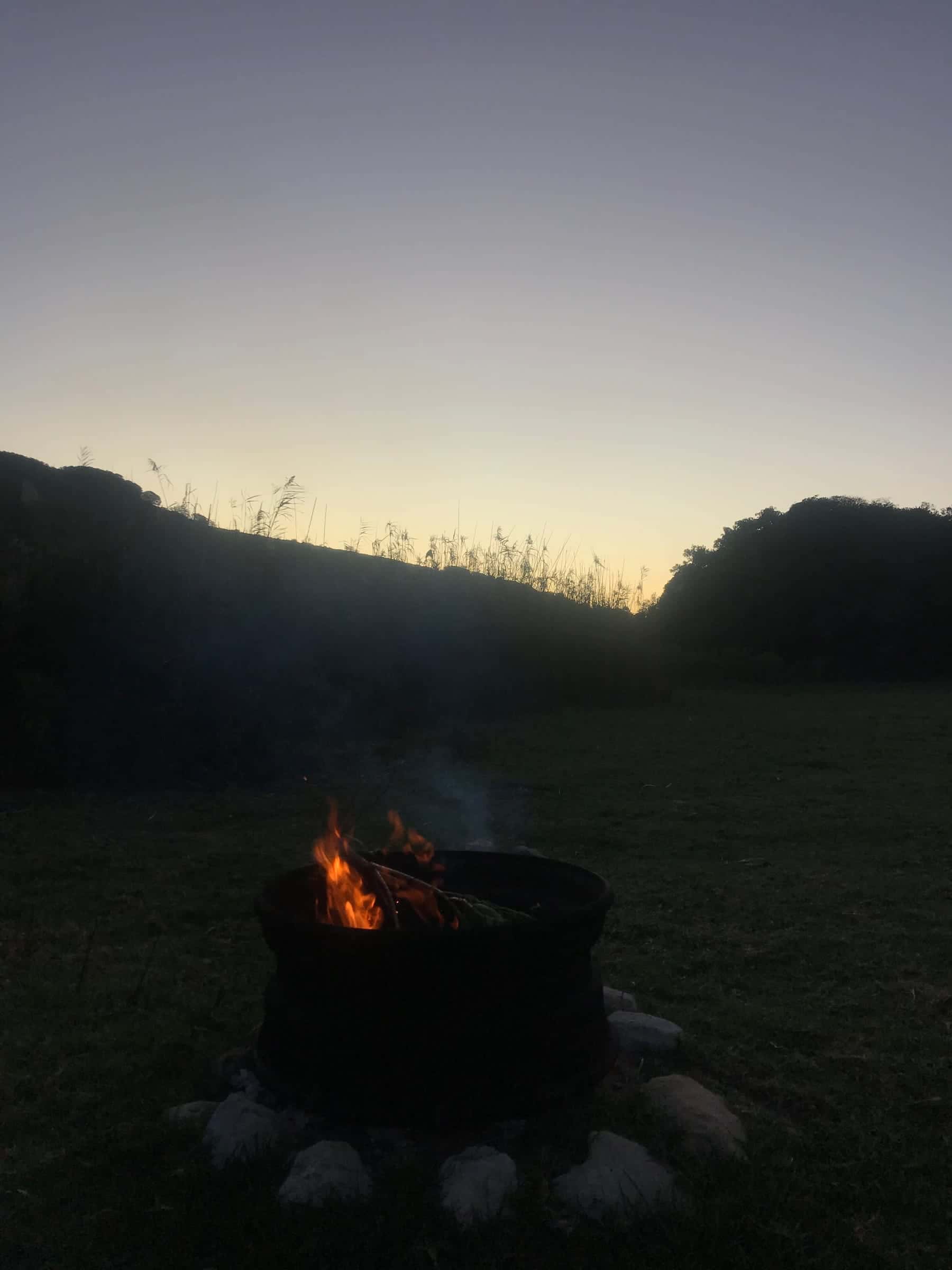 A fire burning in the firepit on the river lawn at dusk, the wooded hills dark behind.