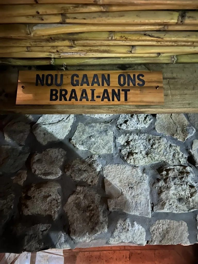 A wooden sign reading "Nou gaan ons braai-ant" above the stone braai.
