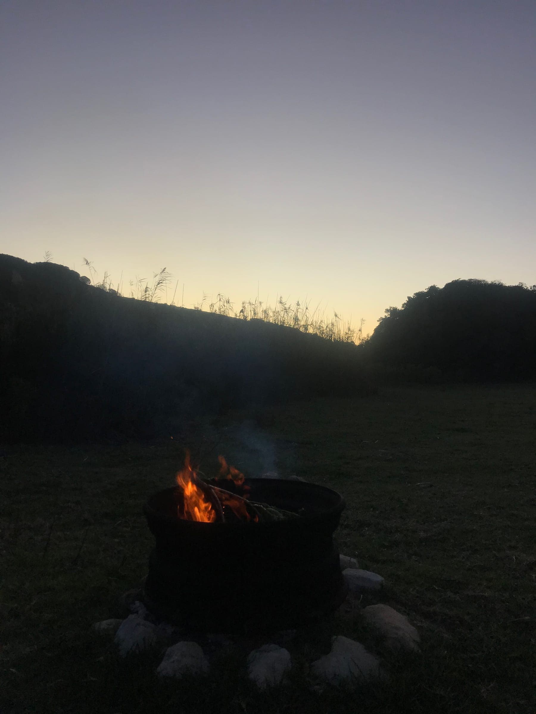 A fire burning in the firepit on the river lawn at dusk, the wooded hills dark behind.