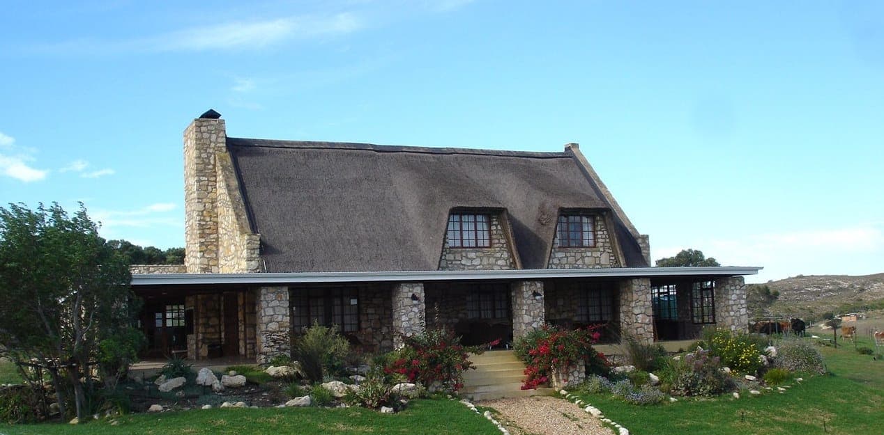 The thatched facade of Stonehouse Lodge near Stilbaai, with its tall stone chimney and dormer windows above the garden.