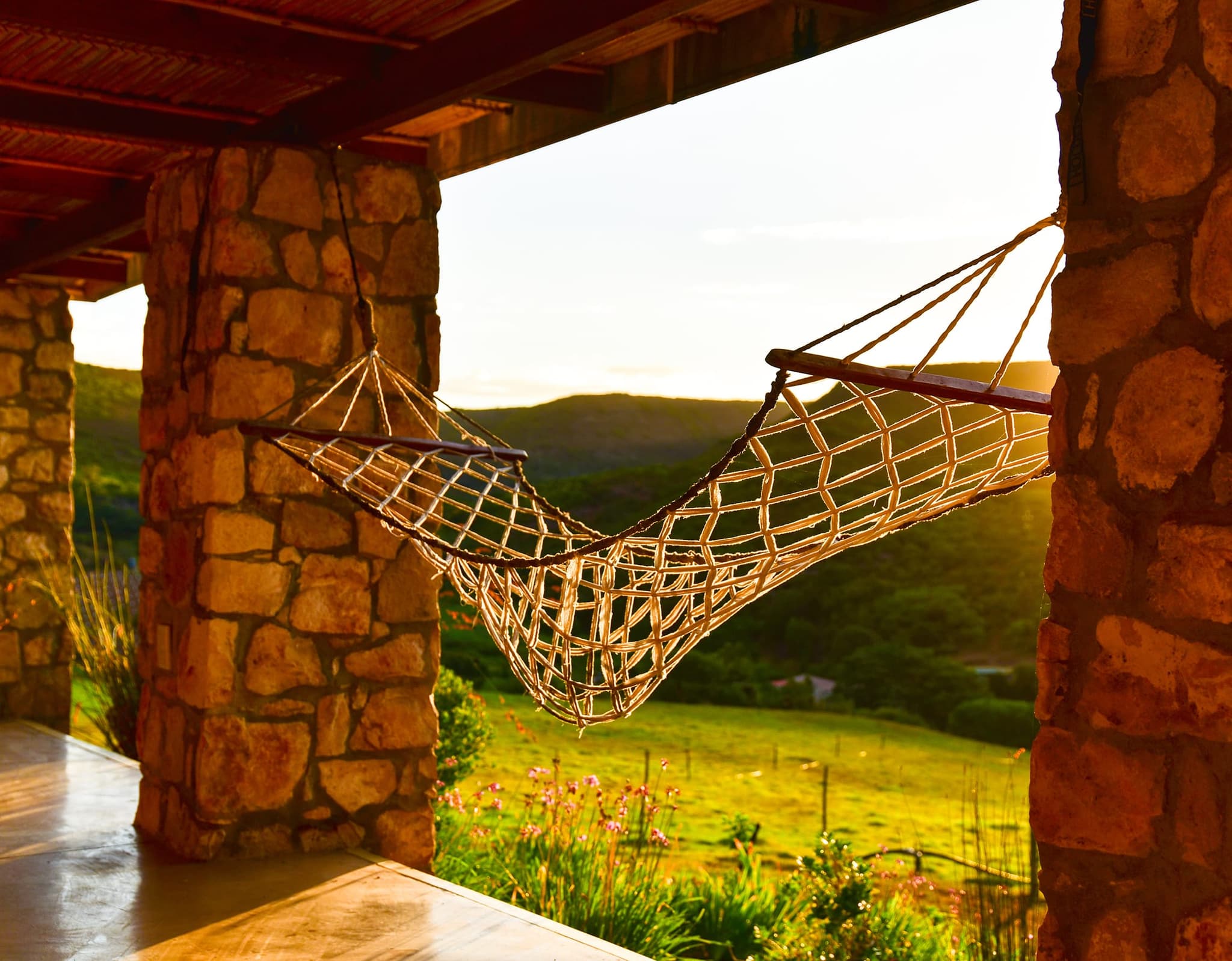 A rope hammock slung between the stone pillars of the Stonehouse Lodge veranda, with green hills and late sun beyond.