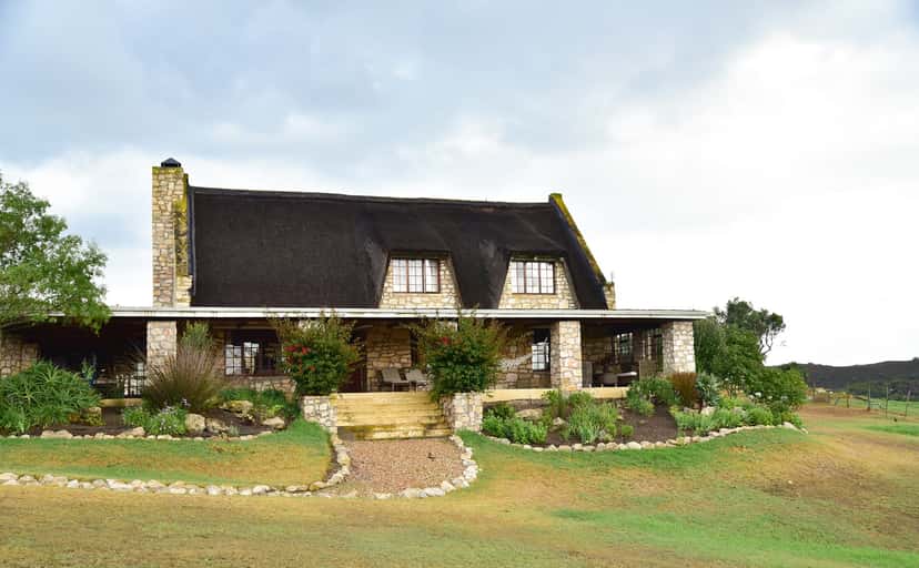 Stonehouse Lodge from the lawn: a thatched stone house with dormer windows, a long covered veranda and a garden path to the steps.